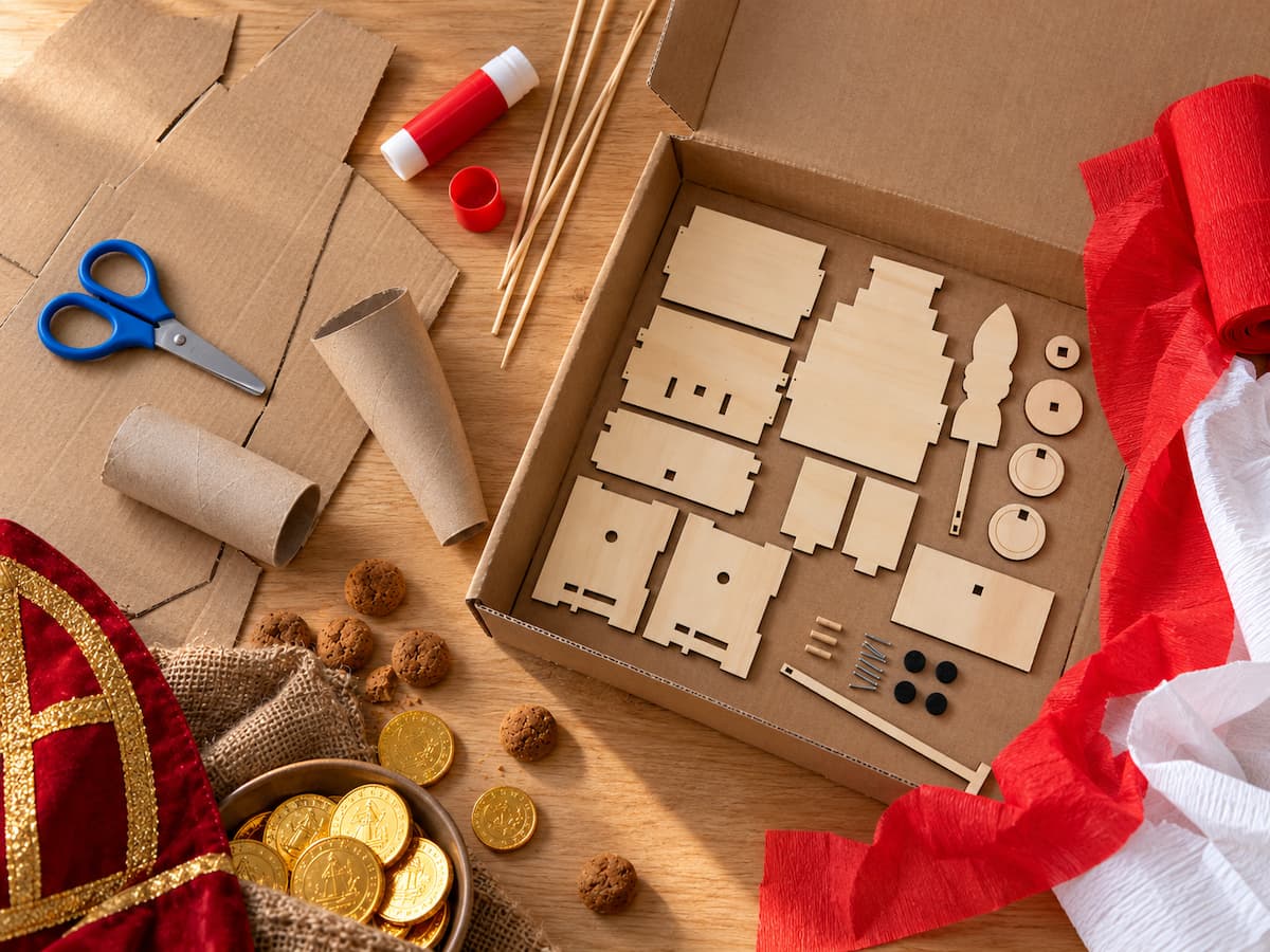 Onderdelen van een houten Sinterklaas-knutselpakket naast een kartonnen doos voor het maken van een surprise, met crêpepapier, een wc-rol en schaar